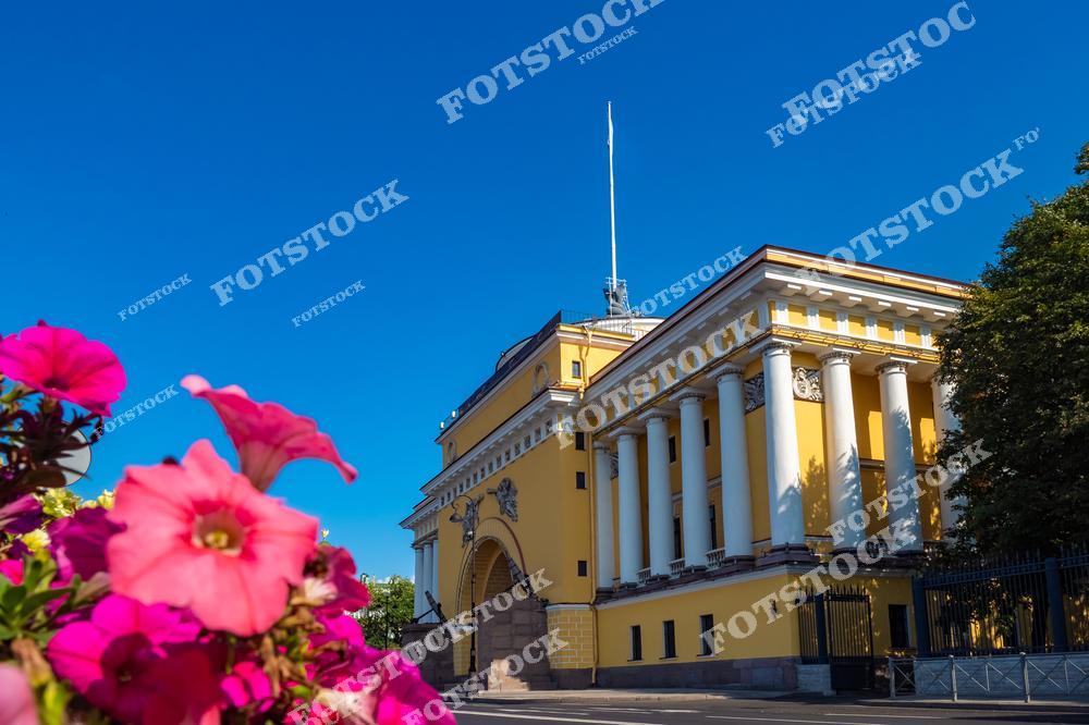 На снимке изображено здание Адмиралтейства в Санкт-Петербурге, выполненное в классическом стиле. Яркие розовые цветы на переднем плане добавляют композиции яркости и контраста. Голубое небо подчеркивает строгую симметрию и величие здания. Адмиралтейство является символом морской истории города и одной из его главных достопримечательностей.